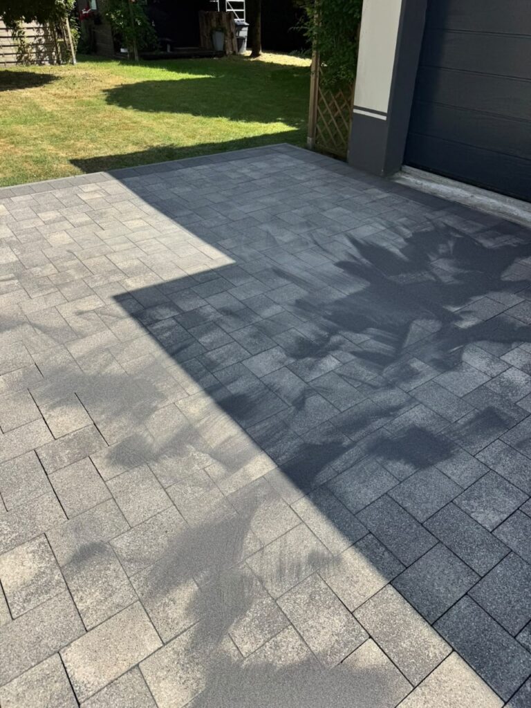 Grey interlocking concrete pavers forming a driveway with strong shadows from a nearby building; green lawn visible to the left and a dark garage door on the right.