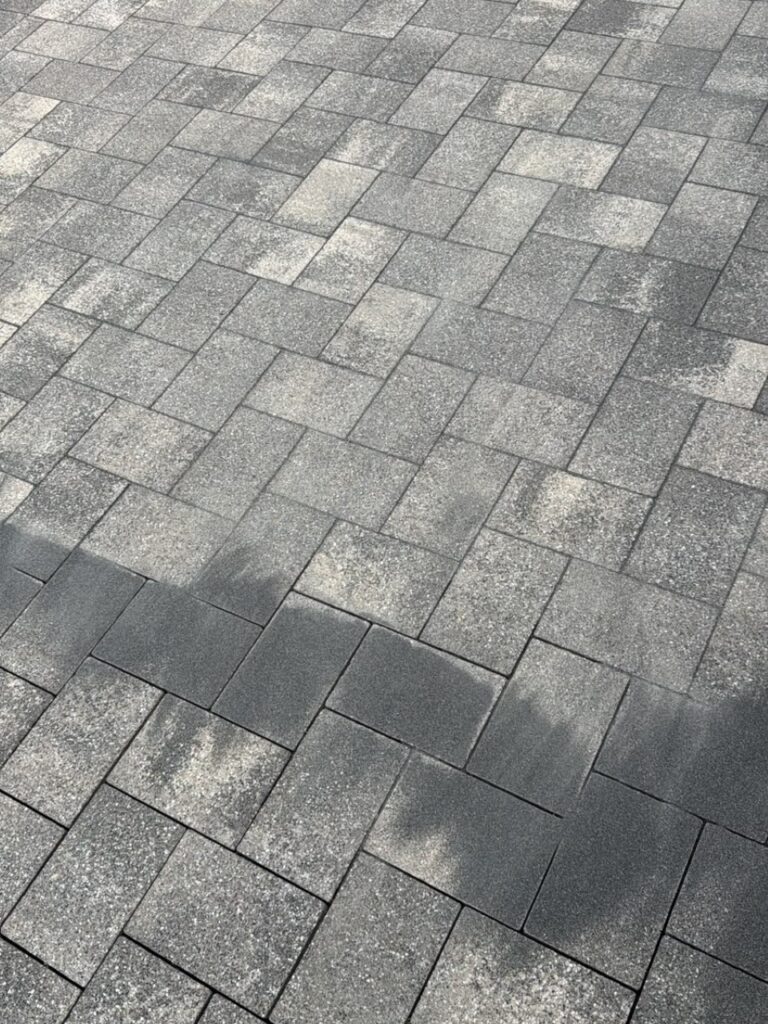 Grey interlocking concrete pavers forming a patterned sidewalk surface with light wear and subtle shadows. OrN/A: Close-up of grey square paving stones in a grid, showing weathered patches and light/dark tones.