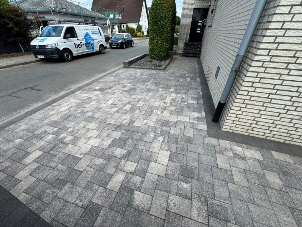 Residential street scene with a gray interlocking-paver driveway beside a white-brick house, a beFresh van and a dark car parked on the road, and a trimmed hedge in a raised bed nearby.