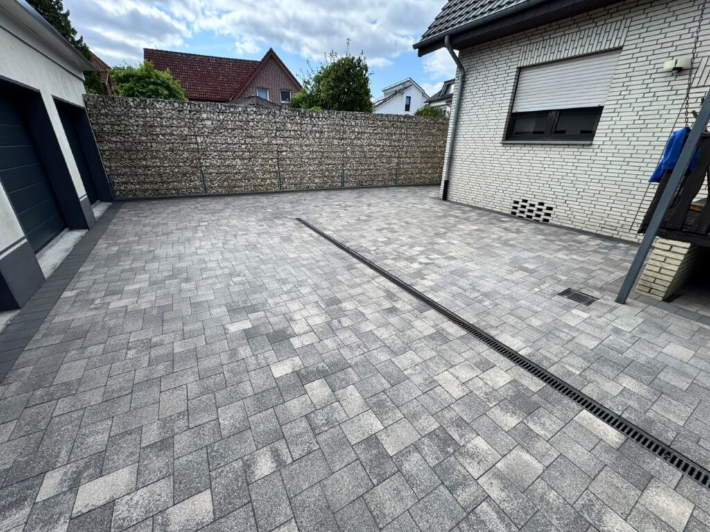 Wide courtyard paved with grey interlocking bricks, a long drainage grate runs diagonally across, with a gabion stone wall at the far end and a white-brick house to the right.